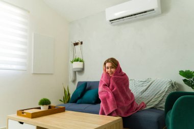 Genç bir kadın battaniyeye sarılmış, arka planda klima sesi yankılanırken evinde üşüyor.