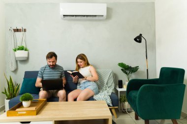 Çift, kanepede dinlenip, kitap okuyup dizüstü bilgisayarla çalışırken klima ünitesinin serinliğinin keyfini çıkarıyor.