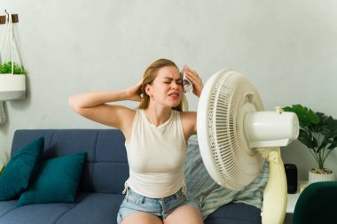 Stresli bir kadın yaz sıcağında serinlemeye çalışıyor. Elektrik vantilatörünün yanında otururken alnından ter siliyor.