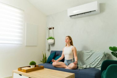 Rahat bir kadın modern dairesinde bir kanepede meditasyon yapıyor, sıcak bir yaz gününde klimanın sağladığı temiz havanın tadını çıkarıyor.