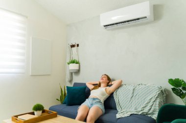 Genç kadın kanepede dinleniyor, mini bölümde serin klimanın keyfini çıkarıyor, yaz sıcağından kurtuluyor.