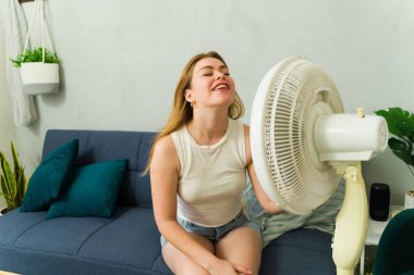Aşırı ısınmış kadın sıcak hava dalgası sırasında evdeki kanepede otururken elektrikli vantilatörden serinletici hava akımının keyfini çıkarıyor.