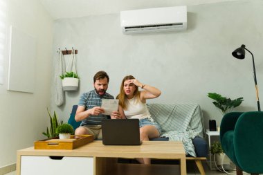 Stresli çift, evdeki kanepede klima açıkken dizüstü bilgisayar kullanarak pahalı elektrik faturalarını kontrol ediyor.