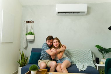 Çift kanepede kucaklaşır, sıcak yaz gününde mini klima ünitesinden serin havanın tadını çıkarır, sıcaktan kurtulur.