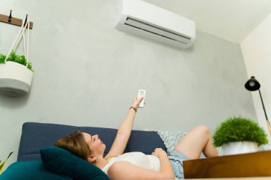 Kanepede uzaktan kumandayla uzanan genç bir kadın sıcak bir yaz gününde klimanın serin havasının tadını çıkarıyor.