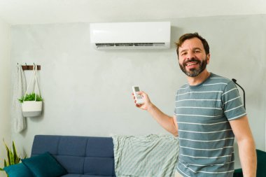 Mutlu bir adam yaz sıcağı sırasında oturma odasında dururken klimayı açmak için uzaktan kumanda kullanıyor.