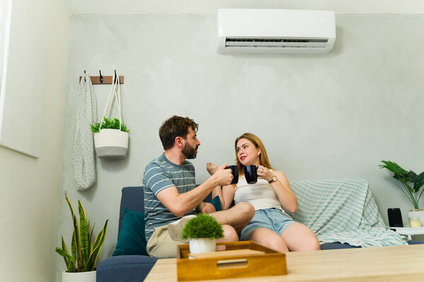 Happy couple relaxing on the sofa at home, enjoying the cool air from their air conditioning unit while sipping beverages