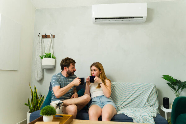 Young couple drinking coffee and talking on sofa under air conditioning unit on hot summer day
