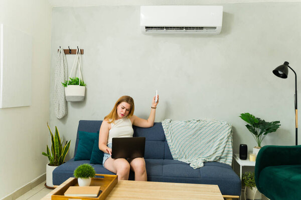 Young woman working at home on her laptop while turning on the air conditioning to cool down during summer heat