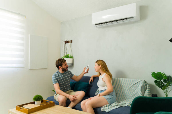 Man pointing air conditioner remote control at woman sitting on sofa, enjoying cool air in living room