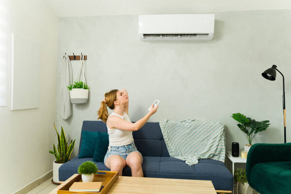 Young woman sitting on sofa using a remote control, adjusting temperature of mini split air conditioner at home