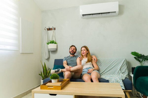 Couple enjoying cool air conditioning while relaxing on sofa in living room and watchint TV during summer heat