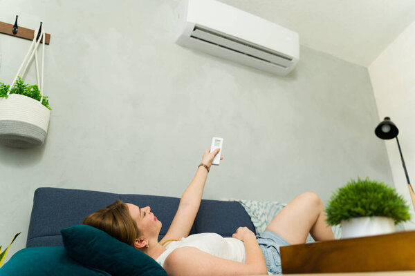 Молодая женщина лежит на диване с дистанционным управлением, наслаждаясь прохладным воздухом из кондиционера в жаркий летний день