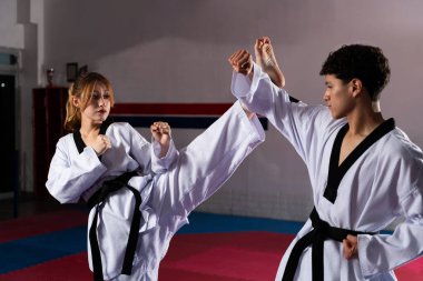 Doboks ve siyah kemer takan iki dövüş sanatçısı bir spor salonunda tekvando tekmesi ve yumruk pratiği yapıyor.