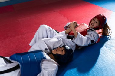 İki tekvando dövüşçüsü antrenman minderinde antrenman yaparken bir kadın, erkek rakibine kol çıtasını indiriyor.