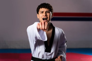 Erkek atlet tekvando çalışıyor, dobok ve siyah kuşak takıyor, gözlemciye yumruk atıyor ve spor salonunun içinde bağırıyor.