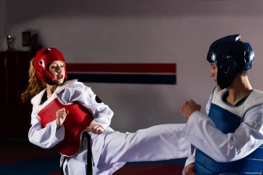 İki sporcu tekvando üniformaları ve koruyucu giysiler içinde idman yaparken, kadın erkeğe güçlü bir tekme atıyor.
