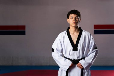 Kendine güvenen dövüş sanatları ustası spor salonunda dikiliyor. Dobok ve siyah kuşak giyiyor. Tekvando eğitimine hazır.