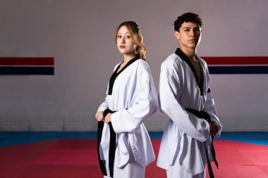 İki genç yetişkin tekvando dövüşçüsü sırt sırta verip sırt sırta verip antrenman minderinin üzerinde dururlar.