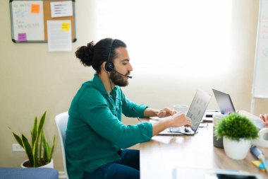 Kulaklık takan Latin işadamı dizüstü bilgisayardaki çizelgeleri analiz ediyor, parlak bir şirkette çalışıyor.