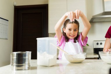 Neşeli genç bir kız, parlak, modern bir mutfakta pişerken kafasına un serpiştirip ortalığı karıştırıyor.