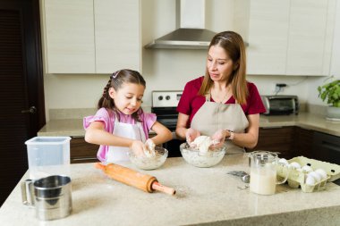 Sevimli bir anne ve kızı hamur yoğuruyor, önlük giyiyor ve modern bir mutfakta yemek pişiriyor.