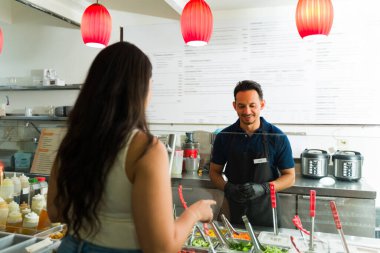 Siyah eldiven giyen Latin restoran çalışanı müşteriye salata barında taze salata sipariş etmede yardım ediyor.