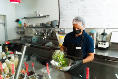 İspanyol aşçı saç filesi takmış ve maske takmış. Ticari bir mutfakta metal bir kasede taze bir salata hazırlıyor.