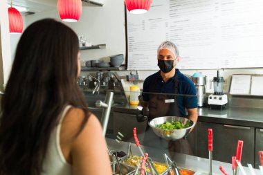 Latin restoran çalışanı yüz maskesi takıp salata barında müşteriye taze salata servis ediyor.