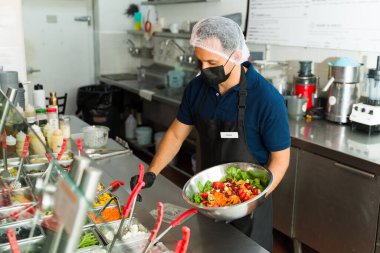 İspanyol şef yüz maskesi takmış ve saç filesi takmış. Restoran salata barındaki müşteriye taze salata hazırlıyor.