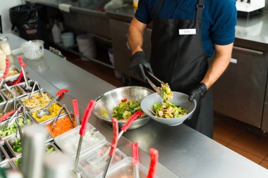 Restoran çalışanı siyah eldiven giyiyor, salata barında maşa kullanan müşteriye taze salata hazırlıyor.