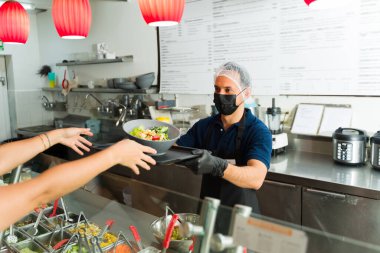 Latin aşçı yüz maskesi ve saç filesi takıp restoran salata barında müşterilere sağlıklı salata tabağı servis ediyor.