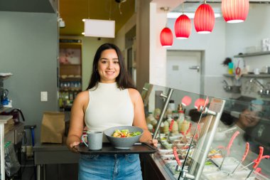 Neşeli bir kadın elinde taze salata ve kahve bardağıyla tepsi tutuyor. Parlak ve modern bir restoranda salata büfesiyle yemeye hazır.