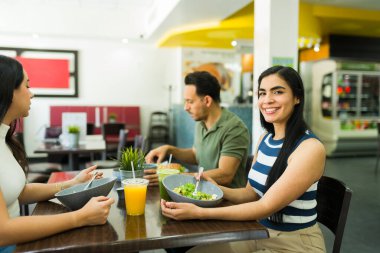 Neşeli genç arkadaşlar canlı bir restoranda taze ve sağlıklı bir öğle yemeği yiyorlar, lezzetli salataların ve içeceklerin tadını çıkarıyorlar.