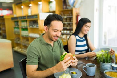 Öğle yemeğinde hamburgerin tadını çıkaran mutlu Latin adam, restoranda bir arkadaşla masada oturuyor.
