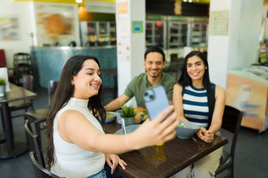 Bir grup mutlu İspanyol genç iş arkadaşı bir restoranda birlikte sağlıklı bir öğle yemeği yerken akıllı telefonlarıyla selfie çekiyor.