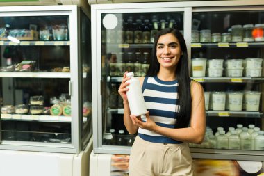 Bir marketin dondurulmuş süt ürünleri reyonunda alışveriş yaparken gülümseyen ve elinde bir şişe kefir tutan genç İspanyol kadın.