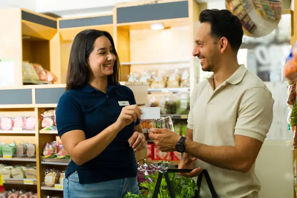 Neşeli süpermarket çalışanı, markette gülümseyip konuşurken erkek müşteriye sadakat kartı veriyor.