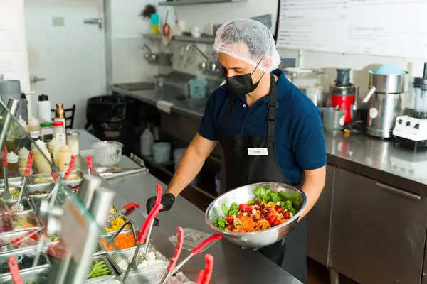 İspanyol şef yüz maskesi takmış ve saç filesi takmış. Restoran salata barındaki müşteriye taze salata hazırlıyor.