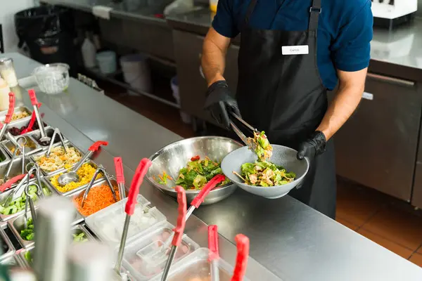 Restoran çalışanı siyah eldiven giyiyor, salata barında maşa kullanan müşteriye taze salata hazırlıyor.