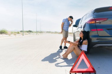 Otoyolda araba kazası geçiren bir çift, yol yardımını aramak için cep telefonu kullanıyor ve bozuk arabalarının yanında bekliyor.