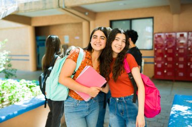 Okul koridorunda sırt çantası ve not defteri taşıyan iki neşeli lise öğrencisi gülümsüyor.