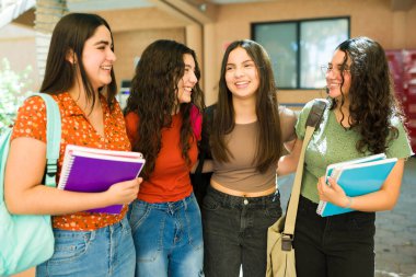 Not defterlerini taşıyan dört mutlu lise öğrencisi okul koridorunda yürüyorlar ve sohbet ediyorlar.