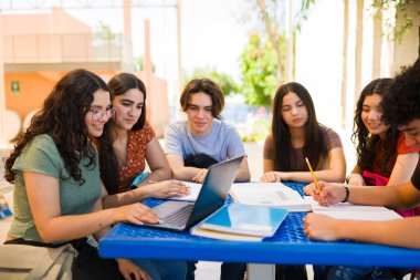 Çeşitli lise öğrencileri bir proje üzerinde birlikte çalışıyorlar, açık bir masada dizüstü bilgisayar ve ders kitapları kullanarak, takım çalışmasını ve modern öğrenmeyi gösteriyorlar.
