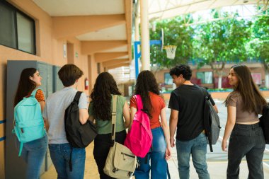 Bir grup farklı lise öğrencisi okul koridorunda birlikte yürüyor, sırt çantaları taşıyor ve dersten sonra sohbet ediyorlar.