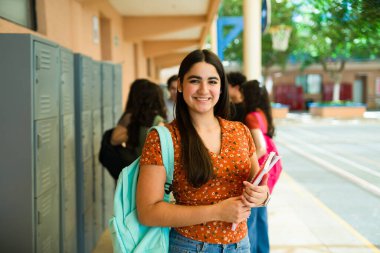 Doldurucu bir koridordaki dolapların yanında gençlik enerjisini somutlaştıran neşeli bir lise öğrencisinin portresi
