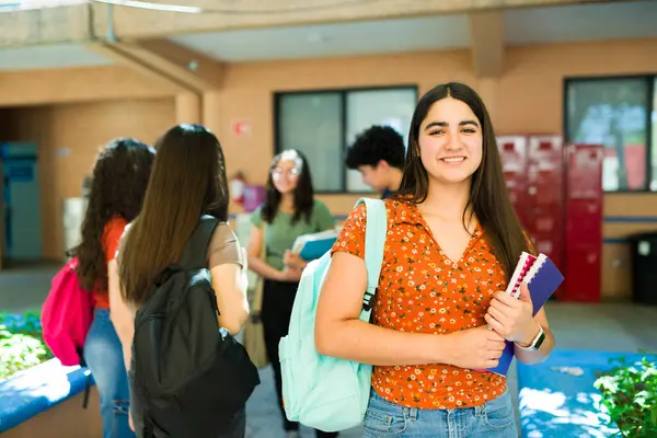 Not defteri tutan ve okul kampüsünde sınıf arkadaşlarıyla dururken sırt çantası takan sevimli bir lise öğrencisi.