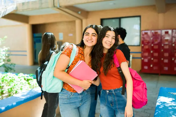 Okul koridorunda sırt çantası ve not defteri taşıyan iki neşeli lise öğrencisi gülümsüyor.