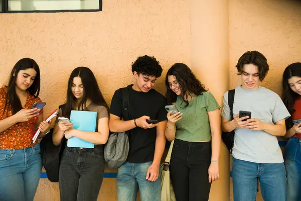 Neşeli Latin lise öğrencileri koridordaki sınıflar arasında akıllı telefonlarını kullanıyorlar.