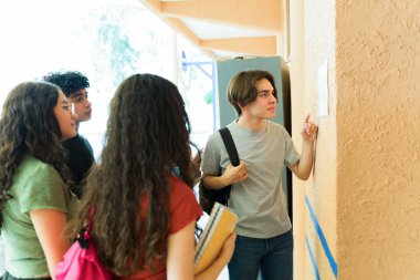 Çeşitli lise öğrencileri, okul koridorundaki bir duvara asılmış önemli bir duyuru okuyorlar.